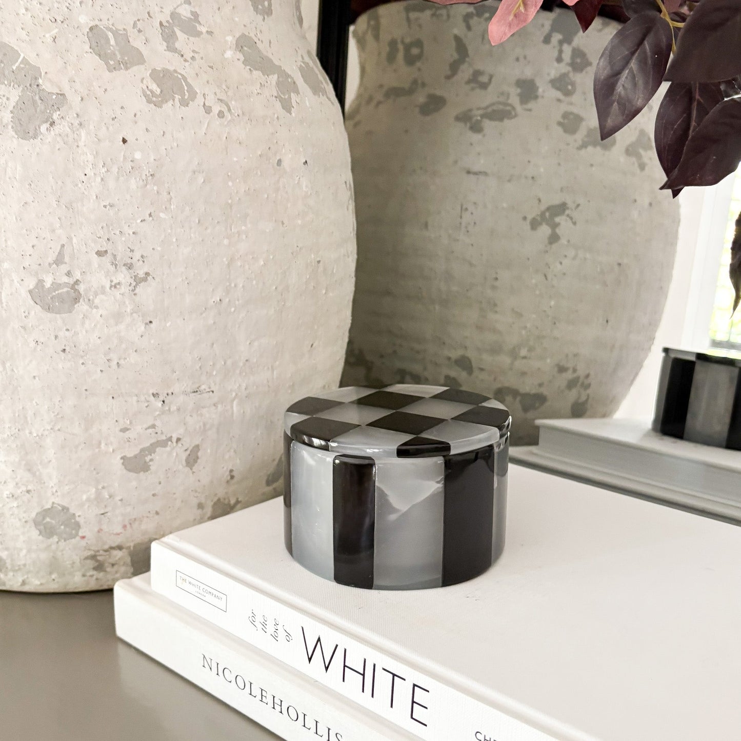 Checkered cylindrical container on a stack of books with decorative vases in the background