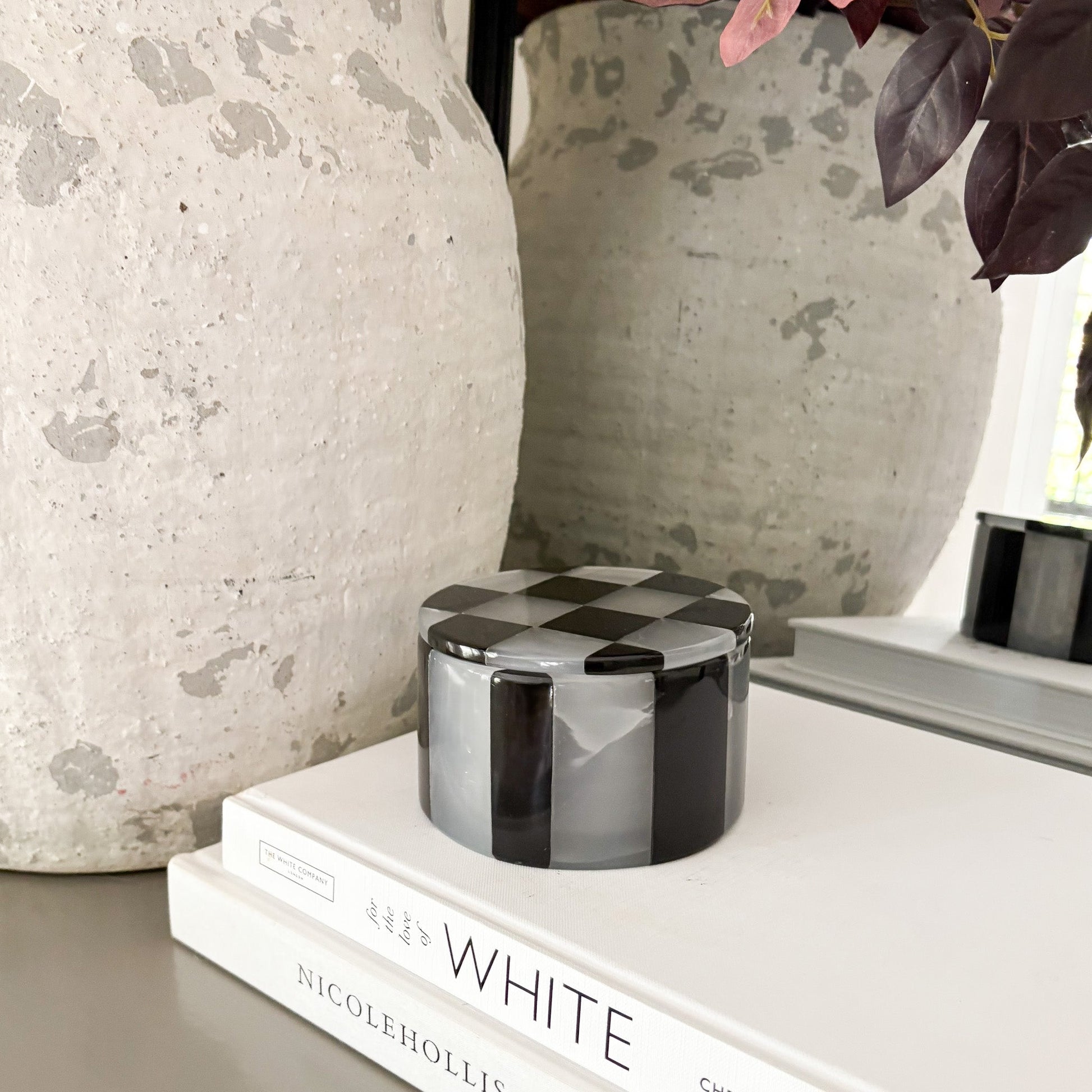 Checkered cylindrical container on a stack of books with decorative vases in the background