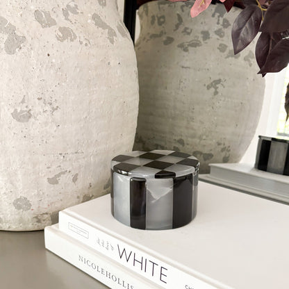 Checkered cylindrical container on a stack of books with decorative vases in the background