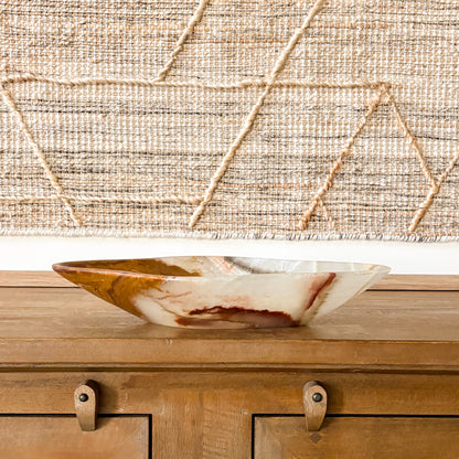 Large hand carved brown onyx bowl with natural veining, displayed on wooden console table with textured wall backdrop