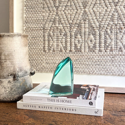 Stack of books with a green glass sculpture on top, next to a textured wall and decorative pot.
