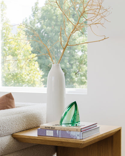 Green glass sculpture displayed on art books beside a white ceramic vase in a bright, modern living room.