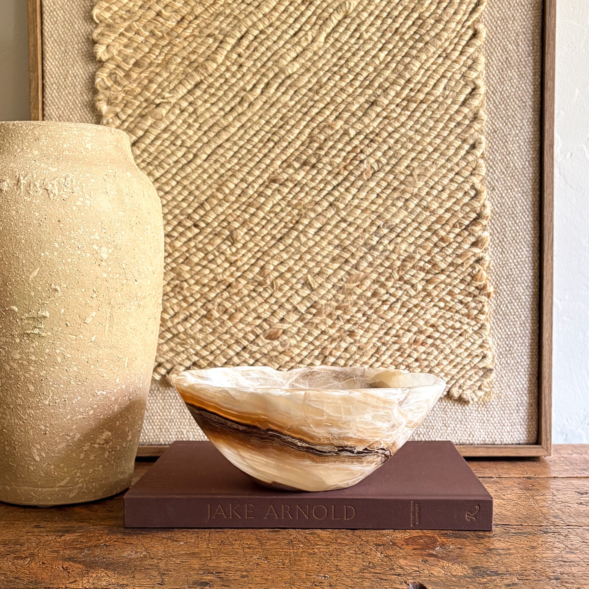 Decorative brown and white onyx bowl on a book with a textured wall and vase in the background