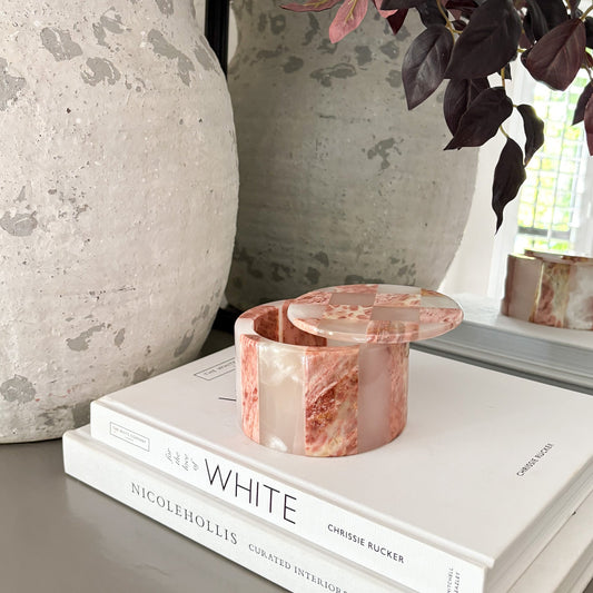 Pink marble trinket dish on a stack of books with decorative vases and a plant in the background.