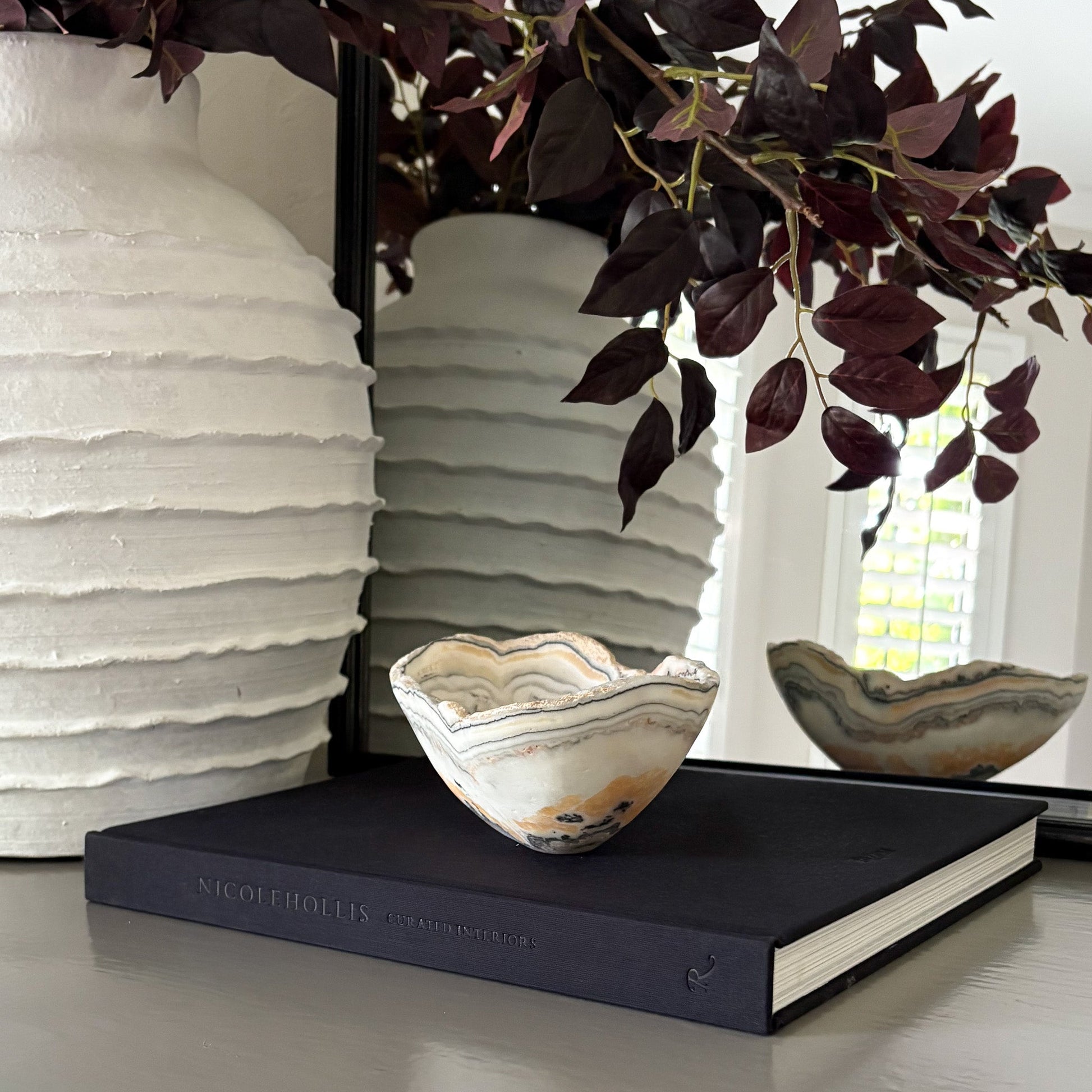 Decorative zebra onyx bowl on a black book with white textured vase and plant in the background