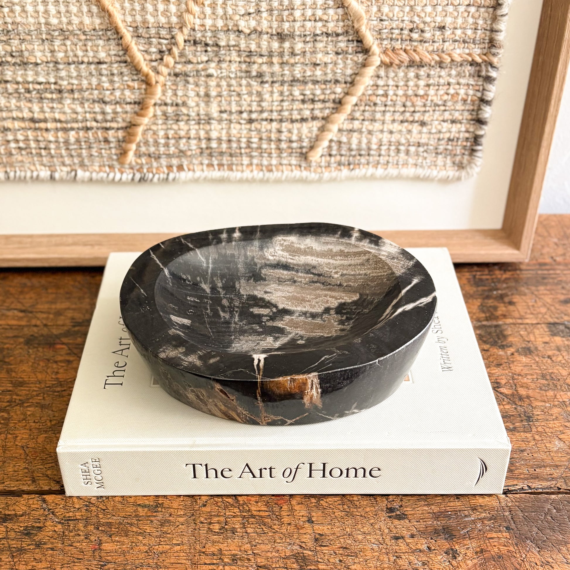 Black fossilized wood bowl on a book titled 'The Art of Home' with a woven wall art in the background.